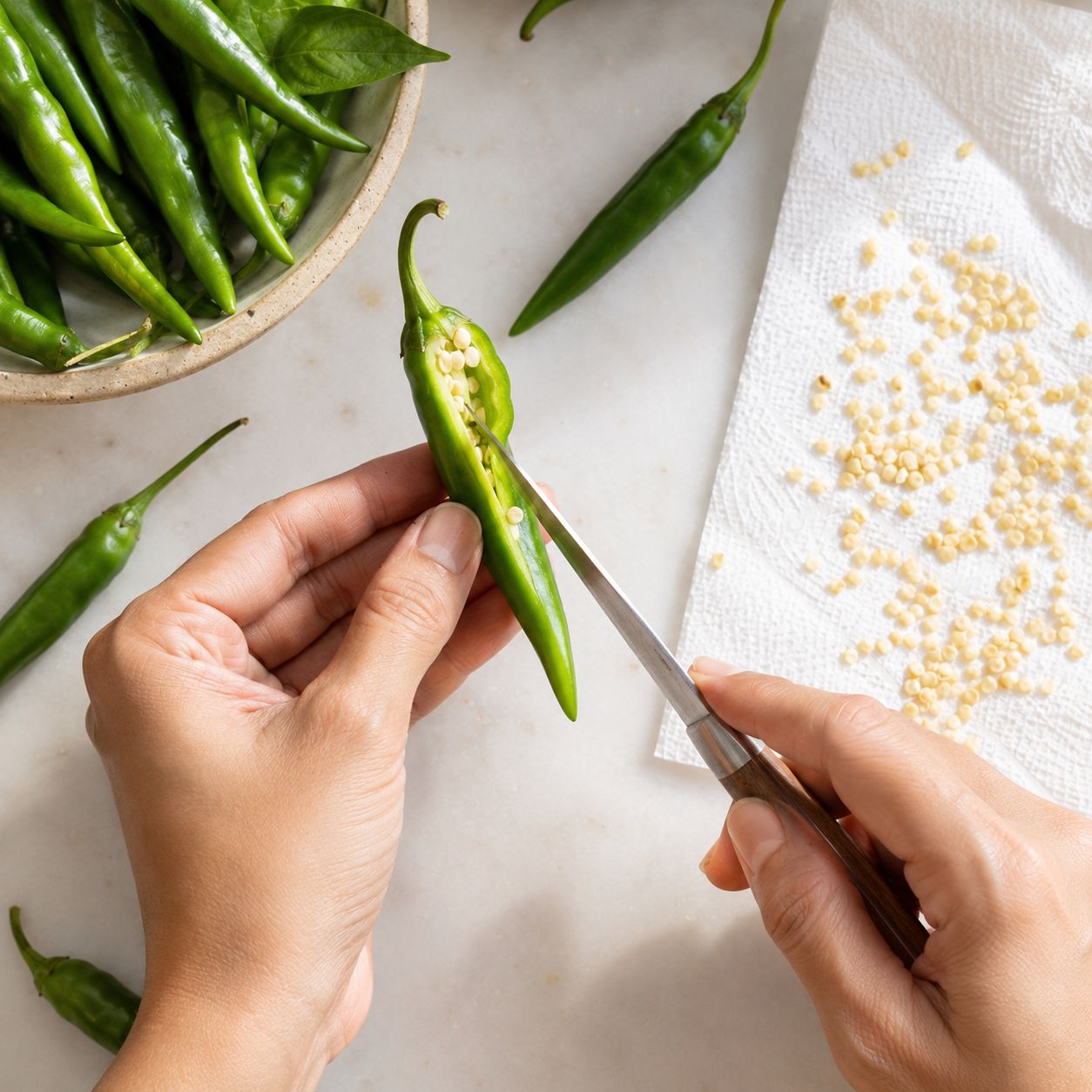 Close-up of hands extracting small pale seeds from a fresh green chilli on a white surface, seeds spread out to dry on paper towel beside it, bright natural light, top-down lifestyle photography