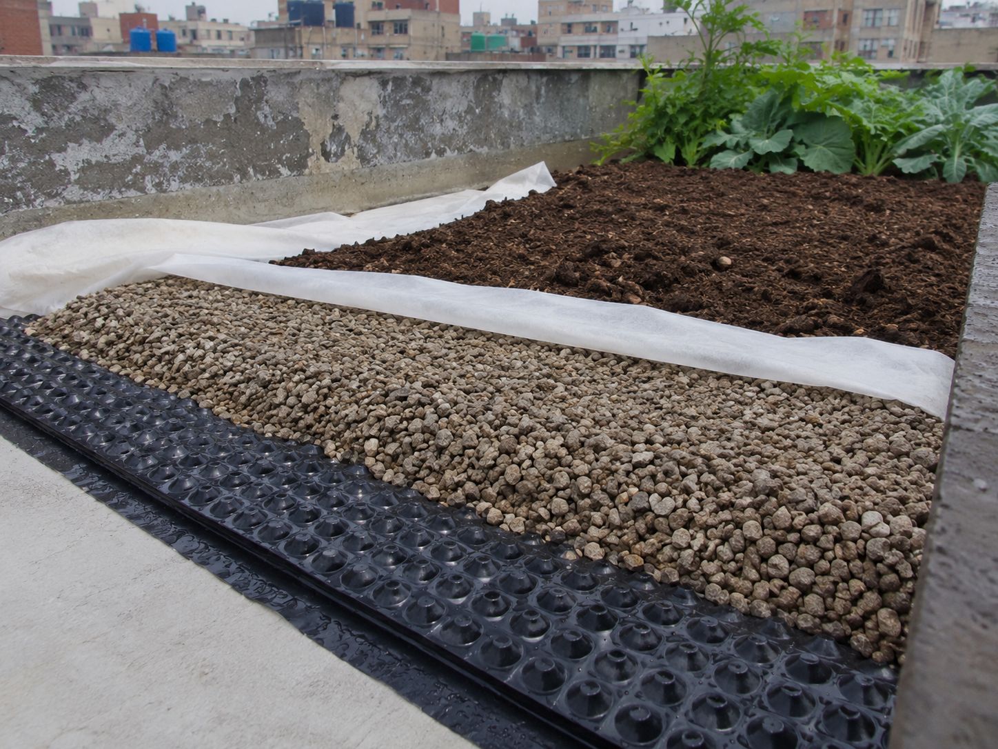 Close-up of a rooftop drainage layer being installed — gravel and drainage membrane laid before the growing medium, showing the layers of a properly waterproofed rooftop garden setup, educational construction photography