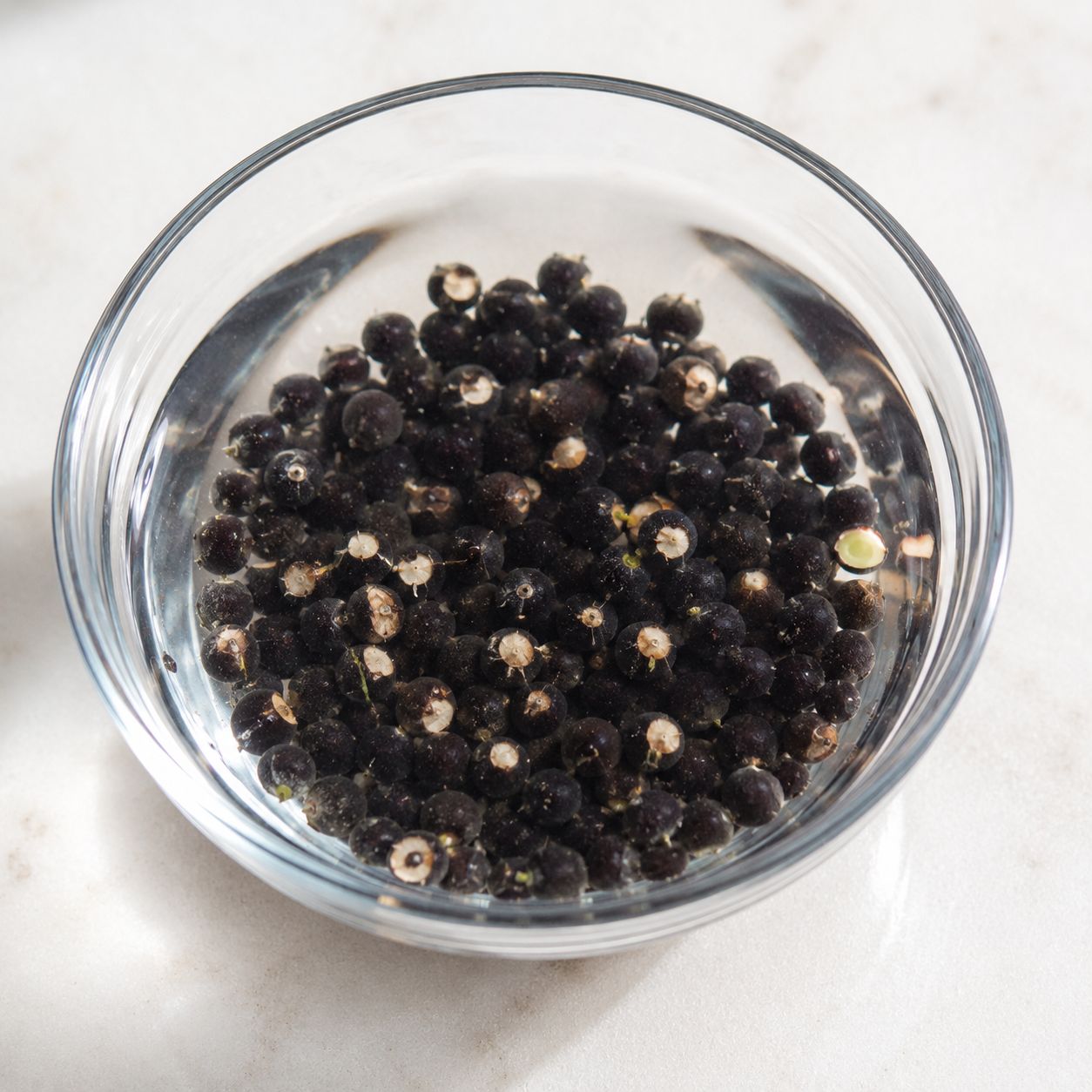 Close-up of okra seeds soaking in a small bowl of warm water overnight on a kitchen counter, the round dark seeds visible beneath the water surface, bright natural light, top-down lifestyle photography